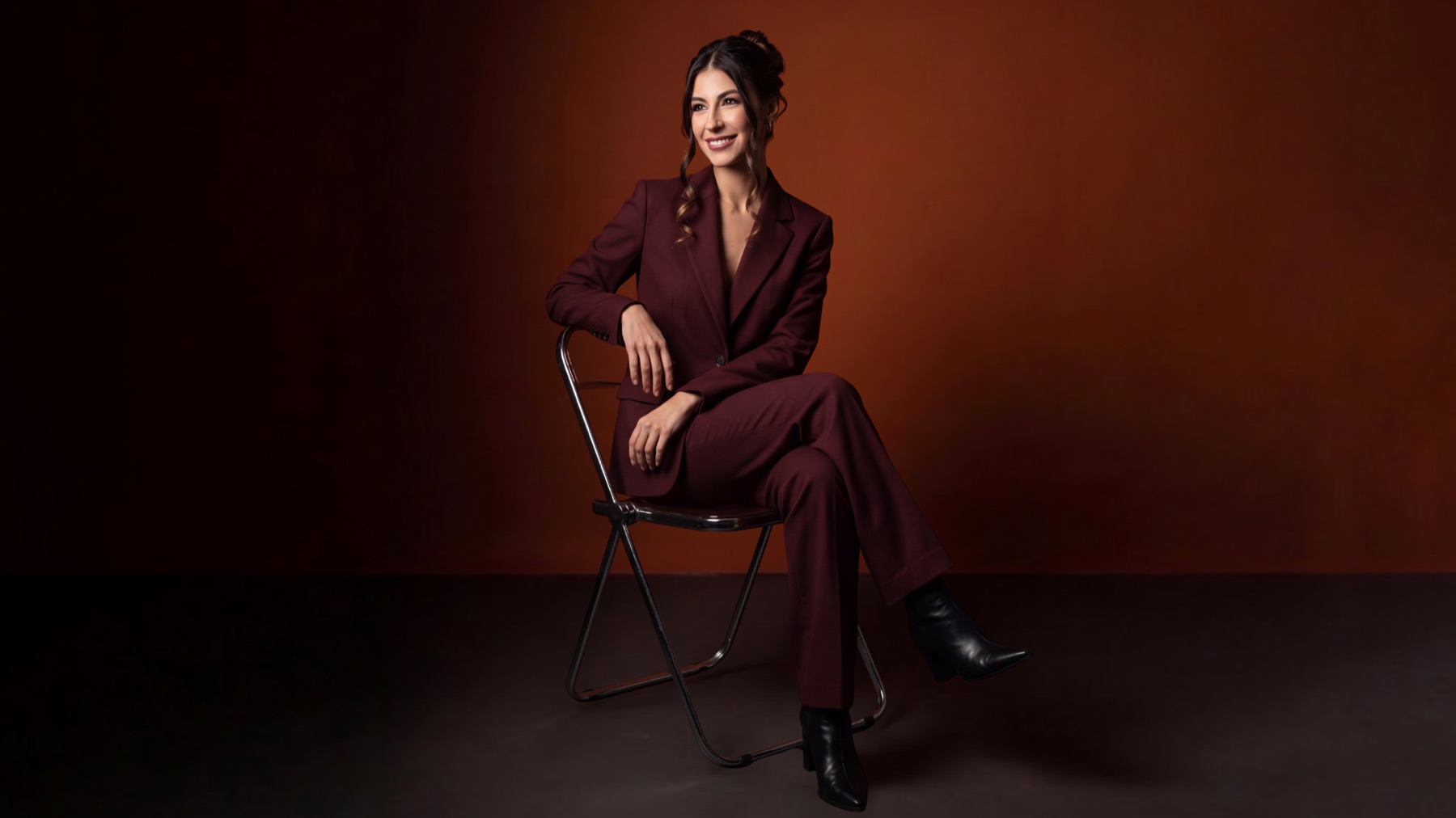 Gabi Corrêa sentada, de terno bordô, sobre um fundo terracota